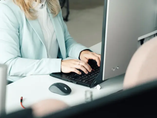 Kollegin im hellen Blazer tippt auf der Tastatur eines Laptops, daneben eine Maus
