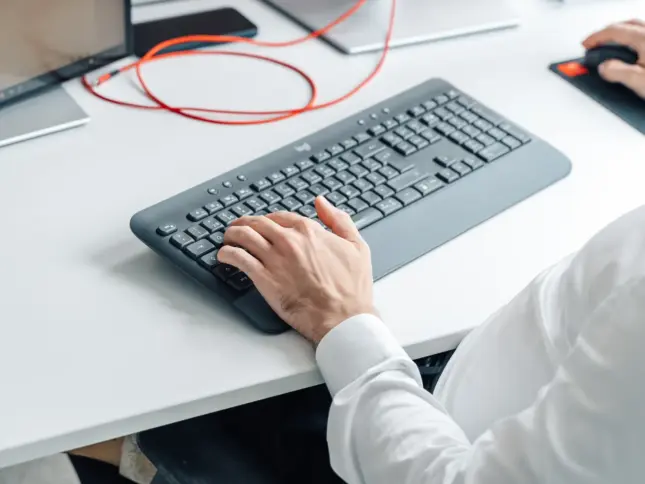 Hand auf einer Tastatur am Schreibtisch, daneben ein rotes Kabel