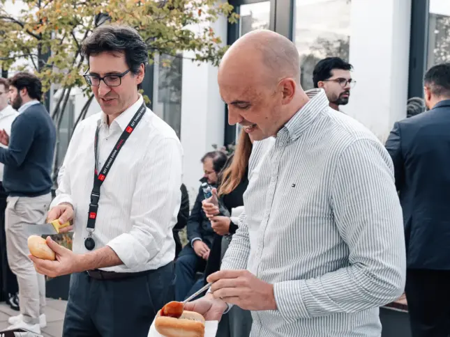 Zwei Kollegen bedienen sich lächelnd am Buffet auf der Dachterrasse, dahinter unterhalten sich Kolleginnen und Kollegen