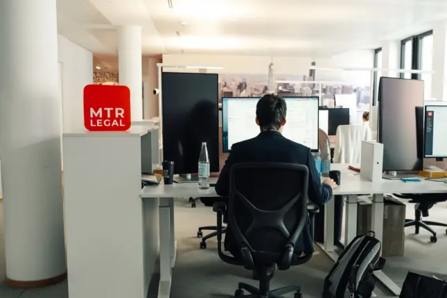 Offener Arbeitsbereich mit Bildschirmplätzen, vorn der rote MTR-LEGAL-Würfel auf einem weißen Schrank